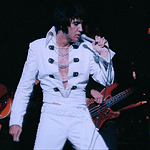 Elvis tribute artist in a white jumpsuit performs on stage, singing into a microphone with a guitarist in the background.