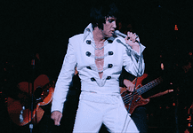 It’s a musical May with music movies all month Elvis tribute artist in a white jumpsuit performs on stage, singing into a microphone with a guitarist in the background.