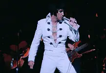 It’s a musical May with music movies all month Elvis tribute artist in a white jumpsuit performs on stage, singing into a microphone with a guitarist in the background.