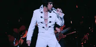 Elvis tribute artist in a white jumpsuit performs on stage, singing into a microphone with a guitarist in the background.