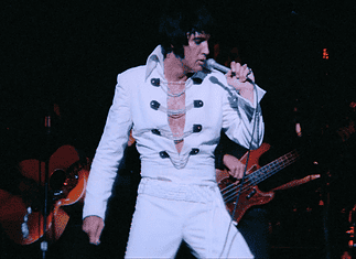 Elvis tribute artist in a white jumpsuit performs on stage, singing into a microphone with a guitarist in the background.