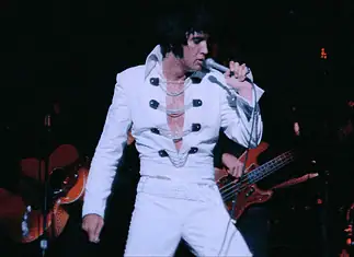 It’s a musical May with music movies all month Elvis tribute artist in a white jumpsuit performs on stage, singing into a microphone with a guitarist in the background.