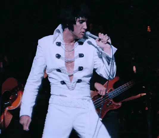 It’s a musical May with music movies all month Elvis tribute artist in a white jumpsuit performs on stage, singing into a microphone with a guitarist in the background.