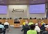 City council chamber with a long wooden dais, officials seated, and a large Edmond logo on the back wall. Two screens read 'Edmond' on the sides. Audience seated in front.