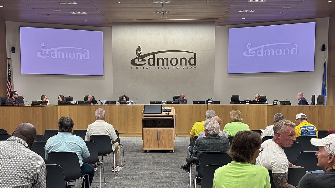 City council chamber with a long wooden dais, officials seated, and a large Edmond logo on the back wall. Two screens read 'Edmond' on the sides. Audience seated in front.