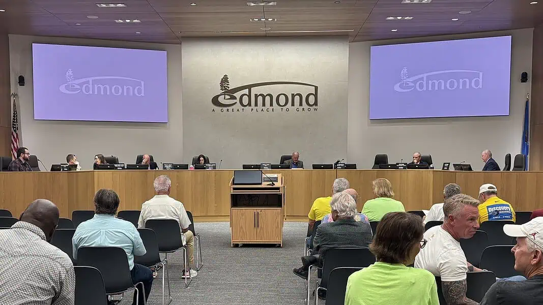 Edmond City Council 5-11-26 City council chamber with a long wooden dais, officials seated, and a large Edmond logo on the back wall. Two screens read 'Edmond' on the sides. Audience seated in front.