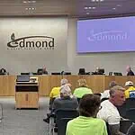 Business as usual at first Edmond City Council meeting since shooting City council chamber with a long wooden dais, officials seated, and a large Edmond logo on the back wall. Two screens read 'Edmond' on the sides. Audience seated in front.