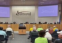 City council chamber with a long wooden dais, officials seated, and a large Edmond logo on the back wall. Two screens read 'Edmond' on the sides. Audience seated in front.