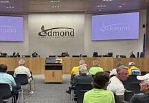 Business as usual at first Edmond City Council meeting since shooting City council chamber with a long wooden dais, officials seated, and a large Edmond logo on the back wall. Two screens read 'Edmond' on the sides. Audience seated in front.