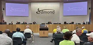 City council chamber with a long wooden dais, officials seated, and a large Edmond logo on the back wall. Two screens read 'Edmond' on the sides. Audience seated in front.