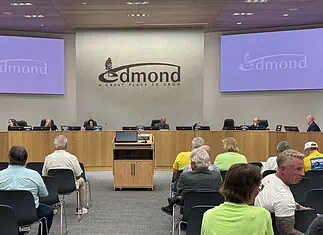 Business as usual at first Edmond City Council meeting since shooting City council chamber with a long wooden dais, officials seated, and a large Edmond logo on the back wall. Two screens read 'Edmond' on the sides. Audience seated in front.