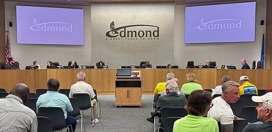 City council chamber with a long wooden dais, officials seated, and a large Edmond logo on the back wall. Two screens read 'Edmond' on the sides. Audience seated in front.