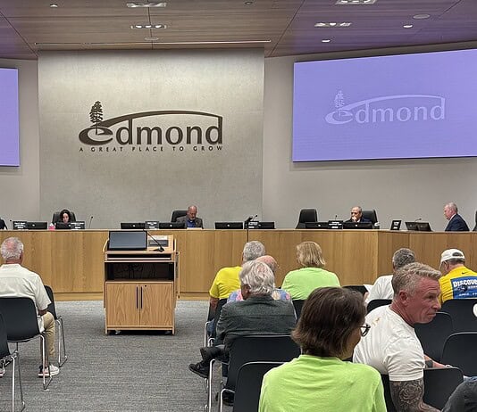 City council chamber with a long wooden dais, officials seated, and a large Edmond logo on the back wall. Two screens read 'Edmond' on the sides. Audience seated in front.