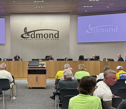 Business as usual at first Edmond City Council meeting since shooting City council chamber with a long wooden dais, officials seated, and a large Edmond logo on the back wall. Two screens read 'Edmond' on the sides. Audience seated in front.