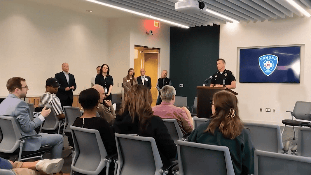 Police officer at a podium addressing a seated audience in a briefing room, Edmond Police logo on a screen behind him.