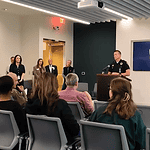 Police officer at a podium addressing a seated audience in a briefing room, Edmond Police logo on a screen behind him.