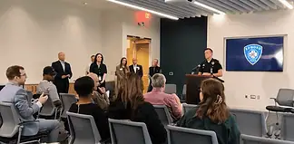 Police officer at a podium addressing a seated audience in a briefing room, Edmond Police logo on a screen behind him.