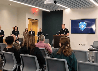 Police officer at a podium addressing a seated audience in a briefing room, Edmond Police logo on a screen behind him.