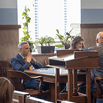 Formal meeting in a boardroom: people seated around a wooden table, a podium in the foreground, plants by the window.