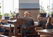 Judge moves Epic Charter Schools embezzlement case forward Formal meeting in a boardroom: people seated around a wooden table, a podium in the foreground, plants by the window.