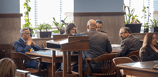Formal meeting in a boardroom: people seated around a wooden table, a podium in the foreground, plants by the window.