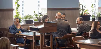 Formal meeting in a boardroom: people seated around a wooden table, a podium in the foreground, plants by the window.