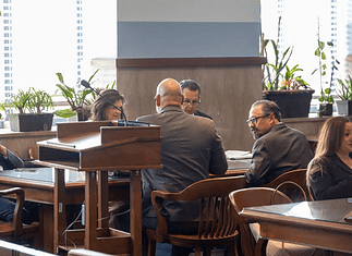 Formal meeting in a boardroom: people seated around a wooden table, a podium in the foreground, plants by the window.