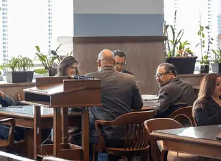 Judge moves Epic Charter Schools embezzlement case forward Formal meeting in a boardroom: people seated around a wooden table, a podium in the foreground, plants by the window.