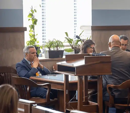 Judge moves Epic Charter Schools embezzlement case forward Formal meeting in a boardroom: people seated around a wooden table, a podium in the foreground, plants by the window.