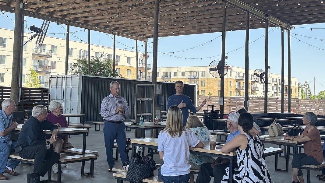 A man in a blue sweater speaks to a small audience seated at picnic tables under an outdoor pavilion, with apartment buildings in the background.