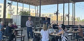 A man in a blue sweater speaks to a small audience seated at picnic tables under an outdoor pavilion, with apartment buildings in the background.