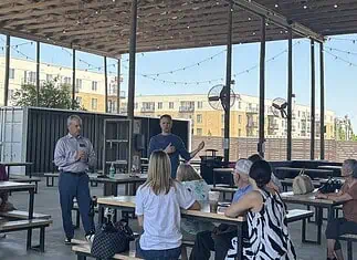 Edmond official says City not investigating police response to shooting A man in a blue sweater speaks to a small audience seated at picnic tables under an outdoor pavilion, with apartment buildings in the background.
