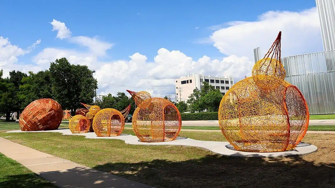 IMG_7604-scaled-e1779896983512 (1) Outdoor art installation featuring large orange wire pumpkins on a curved concrete path in a park under a bright blue sky with white clouds.