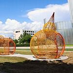 Outdoor art installation featuring large orange wire pumpkins on a curved concrete path in a park under a bright blue sky with white clouds.