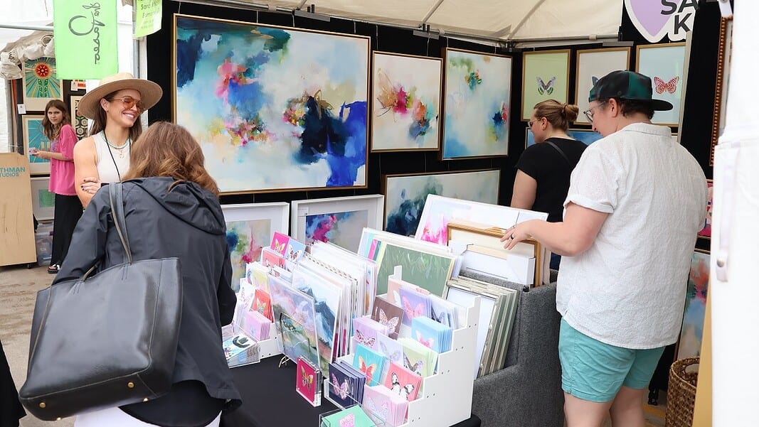 IMG_9453 Art market stall with vivid abstract paintings on the walls and a display of cards and prints; shoppers browse the artwork.