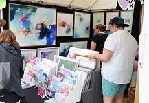 Art market stall with vivid abstract paintings on the walls and a display of cards and prints; shoppers browse the artwork.