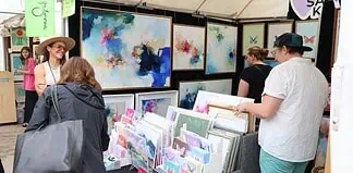 Art market stall with vivid abstract paintings on the walls and a display of cards and prints; shoppers browse the artwork.