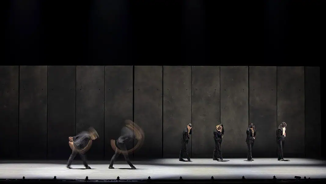 OKCBallet_SHORTS_ANightofPremieres_May2026_JanaCarsonPhoto-17 Line of dancers in black suits on a dim stage, with motion blur from moving bodies behind them.
