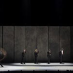 Line of dancers in black suits on a dim stage, with motion blur from moving bodies behind them.