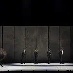 OKC Ballet presents a night of fierce dance, fresh stories with ‘SHORTS’ Line of dancers in black suits on a dim stage, with motion blur from moving bodies behind them.