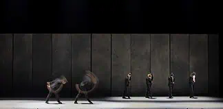 Line of dancers in black suits on a dim stage, with motion blur from moving bodies behind them.