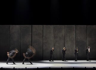 Line of dancers in black suits on a dim stage, with motion blur from moving bodies behind them.