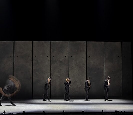 Line of dancers in black suits on a dim stage, with motion blur from moving bodies behind them.