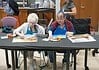 Two women in blue aprons painting at a long table; art supplies and a juice cup are on the table.