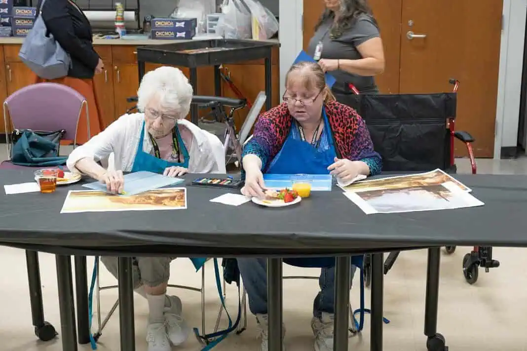 OKCMOA Creative Aging Two women in blue aprons painting at a long table; art supplies and a juice cup are on the table.