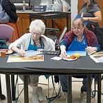 Two women in blue aprons painting at a long table; art supplies and a juice cup are on the table.