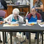 PRESS RELEASE: OKCMOA announces Creative Aging art program for seniors Two women in blue aprons painting at a long table; art supplies and a juice cup are on the table.