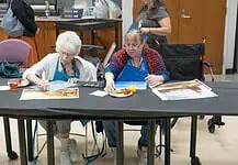 PRESS RELEASE: OKCMOA announces Creative Aging art program for seniors Two women in blue aprons painting at a long table; art supplies and a juice cup are on the table.