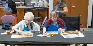 Two women in blue aprons painting at a long table; art supplies and a juice cup are on the table.