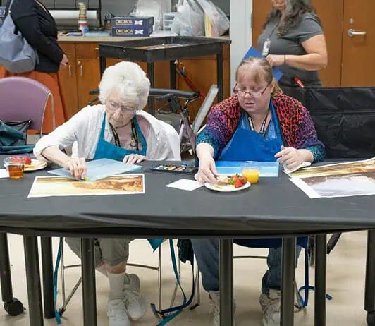 PRESS RELEASE: OKCMOA announces Creative Aging art program for seniors Two women in blue aprons painting at a long table; art supplies and a juice cup are on the table.